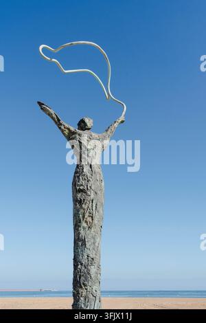 Gijon, Spagna – 19 settembre 2026. Scultura in bronzo solleva le braccia per reggere una fascia bianca curva sopra una spiaggia di sabbia vuota. Lungomare pubblico Foto Stock