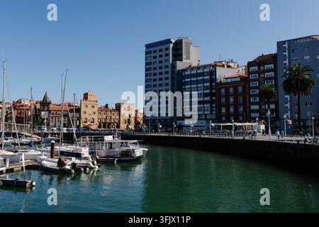 Gijon, Spagna - 19 settembre 2026 Barche a vela sono ormeggiate nella marina sotto il Palacio de Revillagigedo mentre le persone passeggiano lungo la passeggiata sul lungomare. Svago marino Foto Stock