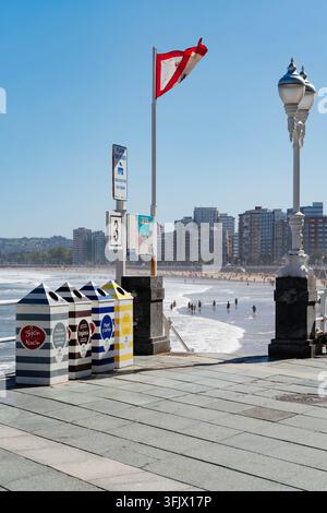 Gijon, Spagna – 19 settembre 2026 Contenitori di riciclo a strisce EMULSA allineano la passeggiata di San Lorenzo mentre i bagnanti si immergono nelle onde. Rifiuti municipali Foto Stock