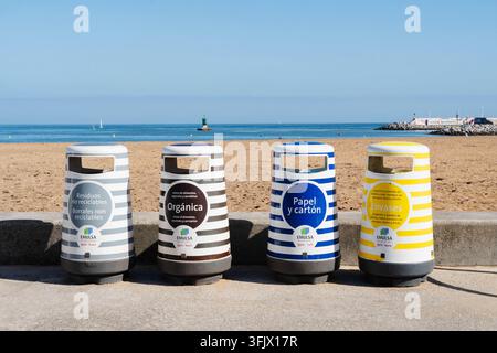 Gijon, Spagna – 19 settembre 2026 Contenitori di riciclo a strisce EMULSA allineano la passeggiata di San Lorenzo mentre i bagnanti si immergono nelle onde. Rifiuti municipali Foto Stock