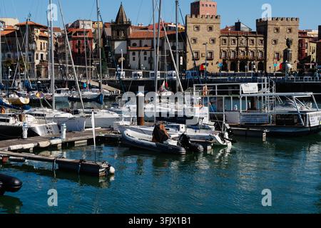 Gijon, Spagna - 19 settembre 2026 Barche a vela sono ormeggiate nella marina sotto il Palacio de Revillagigedo mentre le persone passeggiano lungo la passeggiata sul lungomare. Svago marino Foto Stock