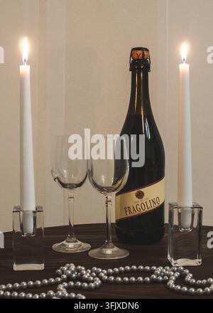 Una bottiglia di spumante Fragolino è appoggiata su un tavolo di legno accanto a due bicchieri trasparenti e candele bianche accese in portacandele di vetro. Foto Stock