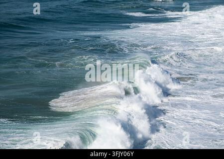 Potente onda oceanica che si infrange sulla superficie del mare aperto, generando schiuma bianca e spruzzi mentre riflette la luce rivelando un delicato arcobaleno nell'acqua Foto Stock