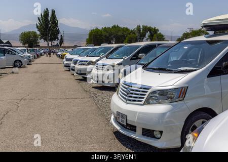 Biscikek, Kirghizistan - 29 giugno 2025: File di minivan sono esposti in un mercato di auto usate all'aperto. Il mercato presenta un lotto pavimentato con montagne in Foto Stock