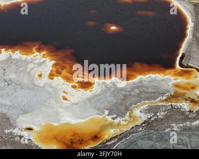 Un'intima ripresa aerea cattura una piscina scura bordata di cristallizzazione arancione ruggine in mezzo a un terreno salino grigio riccamente strutturato. La tavolozza naturale forma una striatura Foto Stock