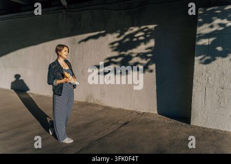Donna sulla quarantina in tuta a righe che cammina lungo un muro di cemento illuminato dal sole tenendo una baguette avvolta nella carta. Foto Stock