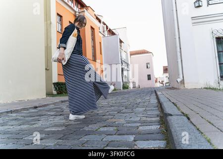 Una donna con una gonna a righe cammina con leggerezza lungo una strada acciottolata colorata, portando una borsa con una baguette dentro. La calda luce del sole, il movimento, un Foto Stock