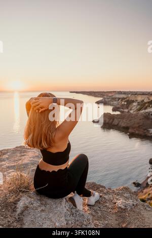 Donna al tramonto seduta su un bordo roccioso che osserva l'ora dorata sul mare rappresentando libertà e pace Foto Stock
