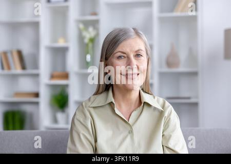 Donna anziana con lunghi capelli grigi che sorride con sicurezza, seduta in un soggiorno moderno e luminoso, rappresentando concetti di invecchiamento elegante, felicità e uno stile di vita confortevole a casa Foto Stock