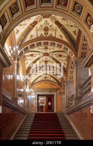 Una larga scalinata rivestita di moquette rossa si innalza sotto una volta riccamente affrescata decorata con grifoni, ghirlande e medaglioni dorati, e candelabri in marmo ornati Foto Stock