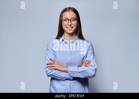 Giovane donna d'affari sorridente con occhiali, in piedi a braccia incrociate, isolata su sfondo grigio Foto Stock