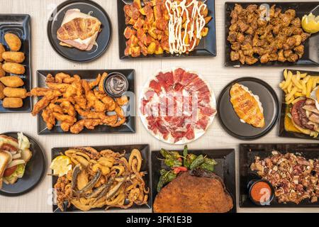 Ampia diffusione aerea di tapas: strisce di pollo, jamón, panini, frutti di mare fritti, burger, ali e cotoletta impanata su piatti neri. Foto Stock