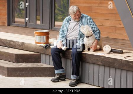 Il proprietario anziano siede con il suo cane su un terrazzo di legno accanto a attrezzature per levigatura e un pennello durante un progetto di restauro all'aperto. Foto Stock