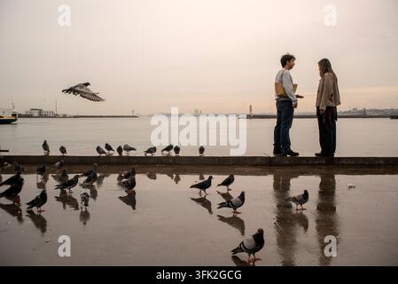 Una giovane coppia si trova insieme sul lungomare di Kadıköy, Istanbul, Turchia, il 16 settembre 2026, circondata da piccioni su una passeggiata bagnata. Foto Stock