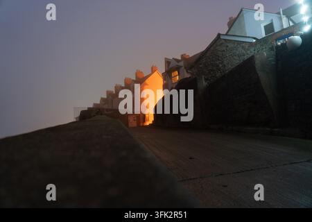 La nebbia serale crea un contrasto suggestivo con l'illuminazione calda delle case su West Street, Watchet, Somerset, catturando il fascino tranquillo della città. Foto Stock