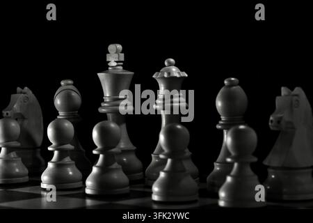 Colpo macro ravvicinato di pezzi di scacchi in legno in piedi su una scacchiera contro uno sfondo nero intenso. L'illuminazione monocromatica drammatica evidenzia le Foto Stock