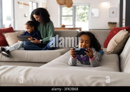 Famiglia afroamericana femminile seduta su divano grigio a casa, usa insieme tablet e smartphone Foto Stock