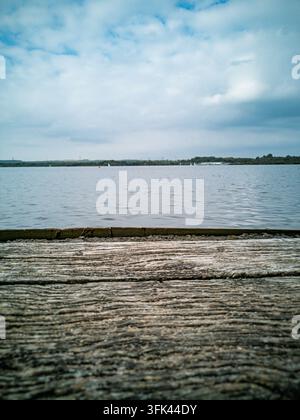Una vista ravvicinata di assi di legno consumate su un molo che conduce a un lago tranquillo con acque calme e rive verdi in lontananza sotto un cielo nuvoloso di giorno Foto Stock