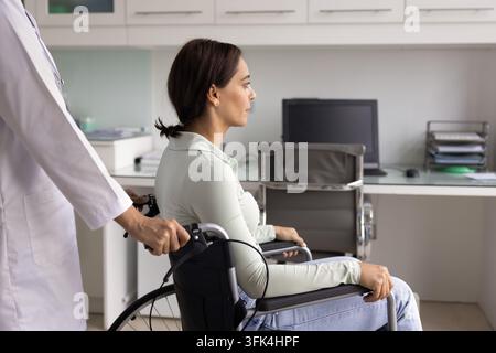 Medica in camice bianco che spinge una giovane donna in sedia a rotelle Foto Stock