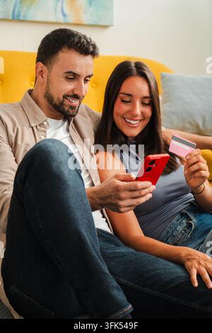 Una coppia sorridente si siede insieme con un telefono e una carta, sfoglia offerte online e prende una decisione condivisa sugli acquisti in un momento confortevole a casa Foto Stock