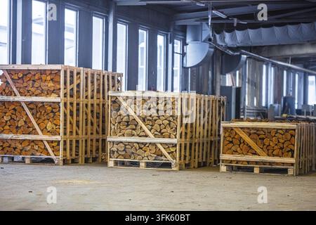 Vista di casse di legno piene di legna da ardere tagliata impilate all'interno di un ampio magazzino industriale con grandi finestre in Lettonia. Foto Stock