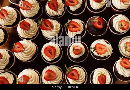 Vista aerea di cupcake al cioccolato ricoperti di glassa alla crema a spirale e mezze fragole fresche disposte vicine per un evento o dessert b Foto Stock