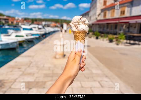 Mano che tiene un cono di gelato su un lungomare soleggiato durante le vacanze estive in Croazia. Godendo dei viaggi mediterranei e dello stile di vita costiero adriatico. Foto Stock