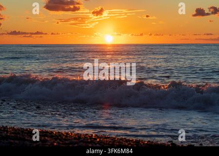 Un'onda del mare che rotola e si infrange sulla riva sotto un vivido tramonto dorato arancione. Foto Stock