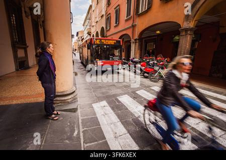 Bologna, Italia – 17 maggio 2018: Vista di una donna in piedi sotto un portico storico mentre un autobus percorre una stretta strada acciottolata con un ciclista che passa Foto Stock