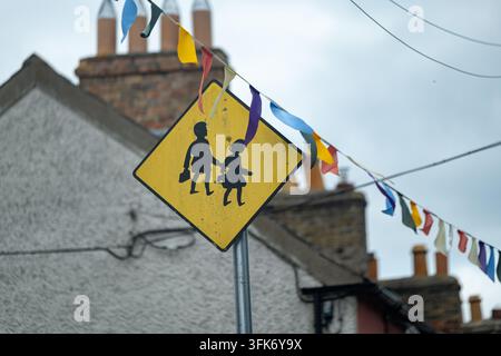Segnale stradale giallo con la scritta "Bambini attraversano la strada" decorato da bandierine colorate, fotografato davanti a una strada residenziale con case sfocate in lontananza Foto Stock