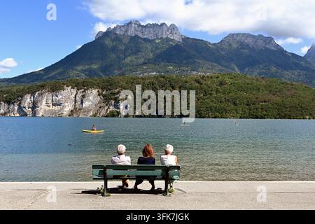 Turisti anziani godono la vista dalle panchine sul lago di Annecy dal lato ovest del lago verso est & il picco delle Dents de Lanfon sopra Talloires Francia Foto Stock