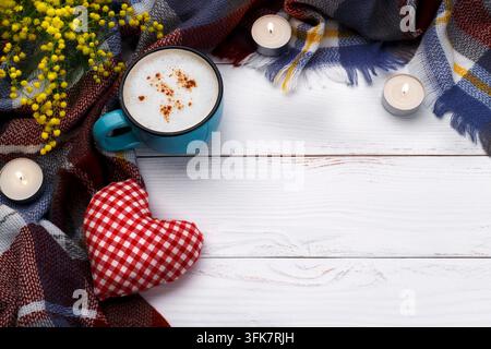 Cappuccino caldo, coperta, candele, fiori di mimosa e cuore di stoffa su sfondo di legno antico. Concetto di relax romantico stagionale, flat lay Foto Stock