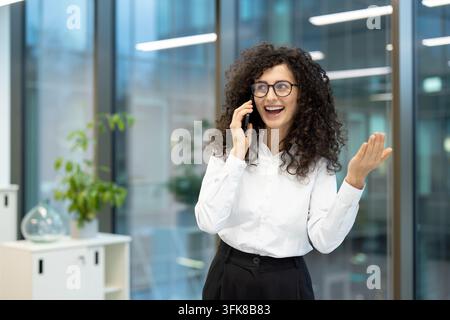Comunicazione aziendale e successo aziendale, donna d'affari felice che parla al telefono in un ufficio moderno con abbigliamento professionale e occhiali da lavoro. Foto Stock