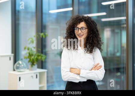 Donna d'affari professionista con capelli ricci in piedi in un ufficio moderno. Manager femminile sicura di sé con occhiali e camicia bianca che guarda lontano con un sorriso mentre lavora in un ambiente aziendale. Foto Stock