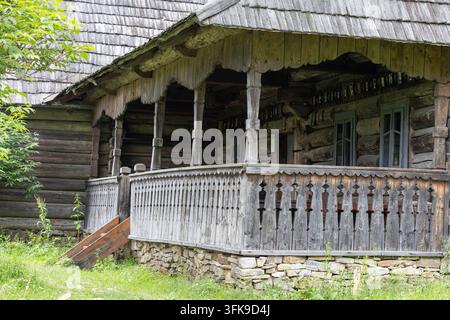 Vista esterna di una vecchia casa in tronchi di legno con tetto a scandole, fondamenta in pietra e portico intagliato in un ambiente naturale. Foto Stock