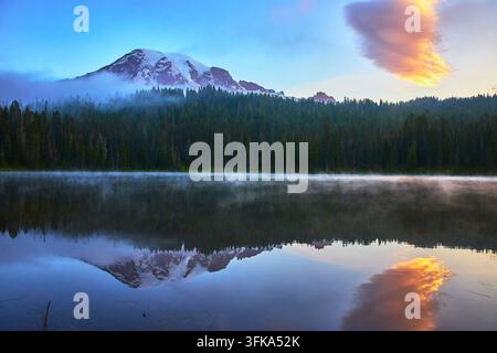La luce dell'alba risplende sul Monte Rainier mentre il lago Reflection riflette la montagna e un cielo vivace. Una leggera nebbia si sposta sull'acqua a Paradise Washingto Foto Stock