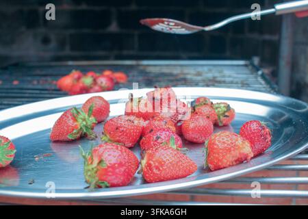 Uno scatto dettagliato di fragole rosse mature disposte su un vassoio d'acciaio con un barbecue e una spatola sullo sfondo. Foto Stock