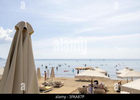 Spiaggia a Zante, Grecia, con ombrelloni chiusi, lettini e turisti che nuotano nel mare calmo con una vasta vista costiera e cielo luminoso durante una Foto Stock