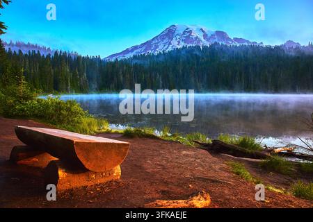 Mount Rainier Riflesso dell'alba sul lago Nebbia e panca di legno in una foresta tranquilla Foto Stock