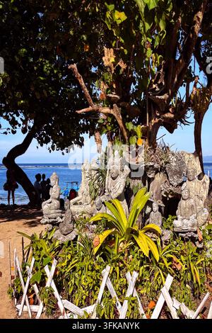 Piccolo santuario da giardino con statue di pietra balinesi sotto un grande albero ombroso sulla spiaggia di Sanur, Bali, Indonesia, 19 aprile 2026. Foto Stock