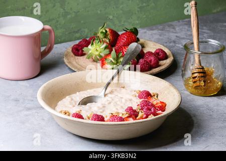 Budino di riso dolce in piatto di ceramica con bacche fragola e lampone, noci, miele e tazza di latte su tavolo da cucina grigio con parete verde come sfondo. Foto Stock