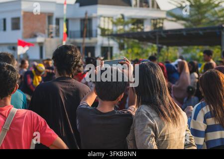 Vista posteriore di una folla diversificata di persone in piedi all'aperto, che guardano e registrano una performance culturale utilizzando i loro smartphone. Foto Stock