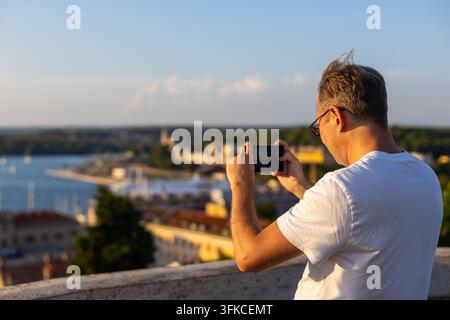 Uomo che fotografa una città costiera con uno smartphone. Uomo che utilizza uno smartphone per fotografare una città costiera e un porto turistico da un punto di vista elevato in luce calda Foto Stock