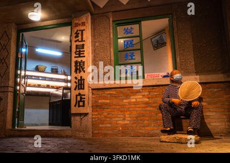 Uomo anziano che si ventola davanti al negozio di cereali e oli Hongxing di proprietà statale su una strada retrò Foto Stock