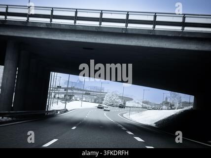Una scena monocromatica vicino all'infrarosso mostra auto che percorrono una strada sotto un sottopassaggio in cemento. La neve copre il terreno sullo sfondo con alberi visibili Foto Stock