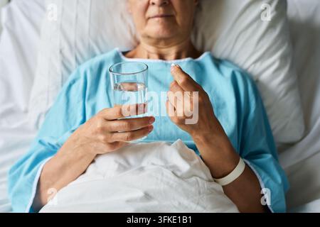 Donna anziana nel letto d'ospedale che tiene una pillola di medicinale e un bicchiere d'acqua Foto Stock