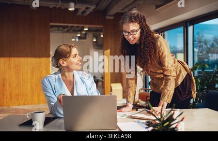 Mentore entusiasta si china sulla scrivania scrivendo appunti mentre la sua collega bionda sorridente alza lo sguardo da un laptop Foto Stock