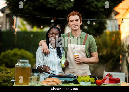 Coppia diversa che prepara il cibo e si gode bevande durante un barbecue all'aperto Foto Stock