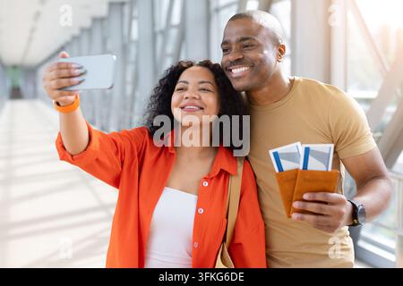 Giovane coppia nera che scatta un allegro selfie con lo smartphone in aeroporto mentre tiene i passaporti Foto Stock