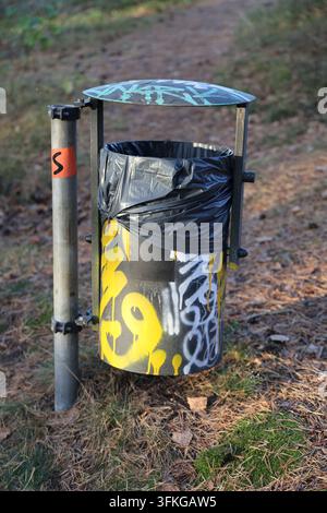 Un bidone della spazzatura pubblico all'aperto coperto di graffiti con un rivestimento in plastica nera, montato su un palo di metallo in un'area boschiva con aghi di pino. Foto Stock